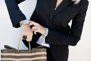 Person in a black blazer holds a striped shopping bag, wearing a silver watch, black ring, and silver bracelets.